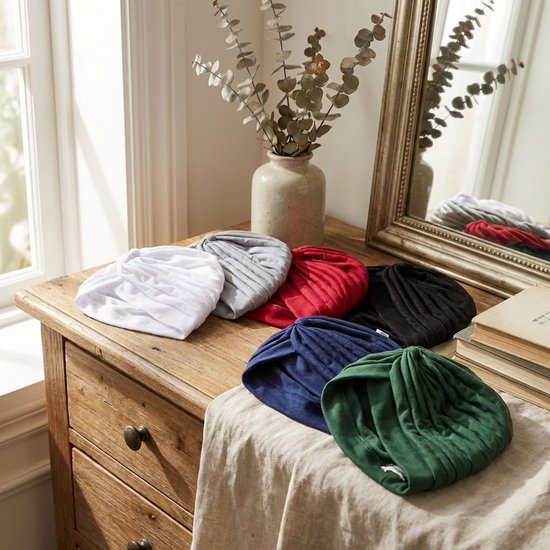 Verschillende tulbanden (wit, grijs, rood, zwart, marineblauw, groen) op een houten commode bij een raam.