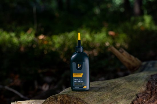 Fles 'All Weather Bio Chain Oil' op een boomstronk in bosrijk gebied.