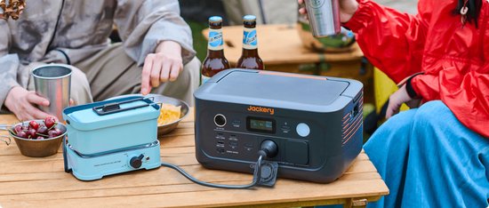 Donkergrijs Jackery Explorer 500 powerstation met oranje accenten, op een houten tafel, aangesloten, met mensen op de achtergrond.