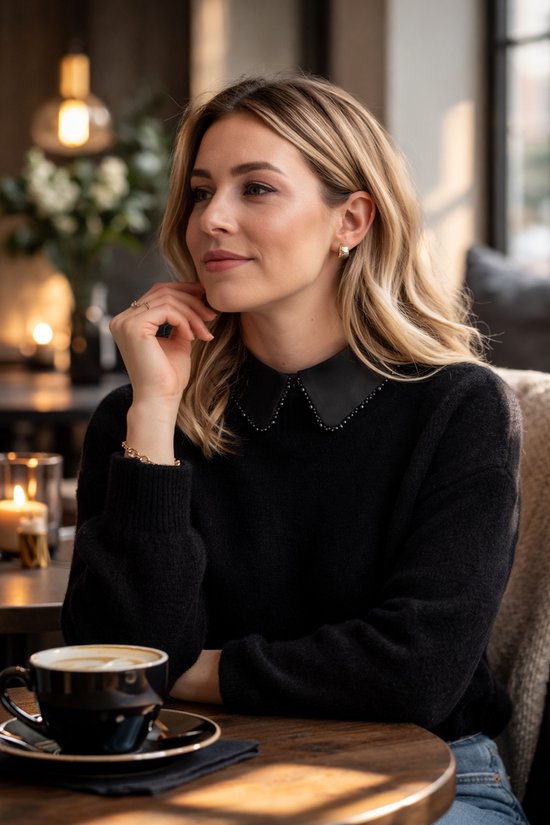 Model met blond haar draagt zwarte trui met kraagje versierd met kraaltjes en gouden oorbellen.