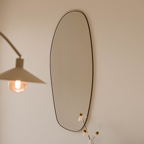 Een organische, zwarte spiegel aan de muur, met een lamp en bloemen ernaast.