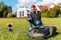 Een jonge vrouw met een smartphone in de tuin bij een robotmaaier en een Netatmo regenmeter.