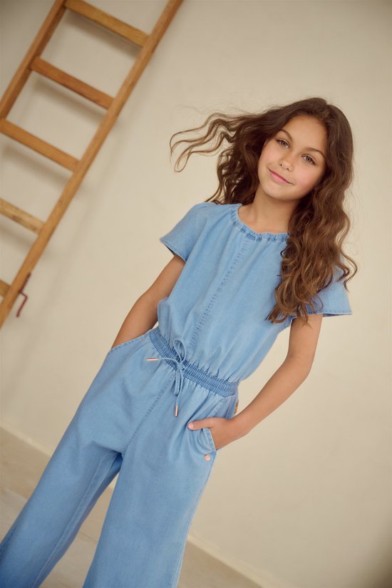 Meisje met lang bruin haar draagt een lichtblauwe denim jumpsuit met korte mouwen en wijde pijpen. Ze staat bij een ladder.