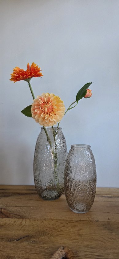 Twee glazen vazen met een ruw, bobbelig oppervlak, op een houten tafel. De onderste vaas bevat oranje en roze bloemen.