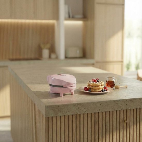 Roze mini wafelijzer op een aanrecht in een moderne keuken, naast wafels met fruit en honing.
