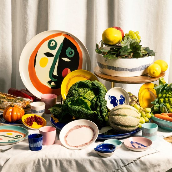 Een gedekte tafel met veel groenten, fruit en verschillende borden, waaronder een grote serveerschaal met een gezicht.