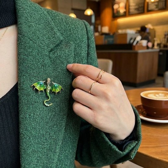 Groene drakenbroche met glinsterende vleugels, gedragen op een groene jas. Gouden ringen op de hand.