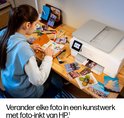 Vrouw met blauwe hoodie werkt aan een schetsboek naast een witte HP printer, met foto's op de houten tafel.