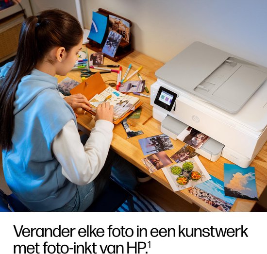 Vrouw met blauwe hoodie werkt aan een schetsboek naast een witte HP printer, met foto's op de houten tafel.