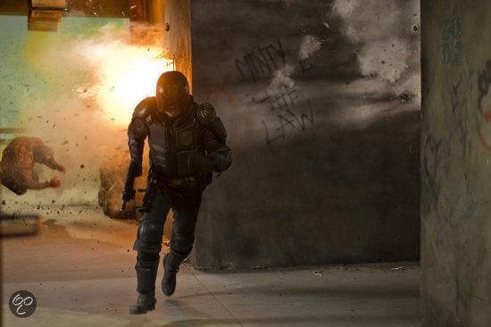 Een figuur in een zwart harnas, rennend met een wapen, met een explosie achter zich.