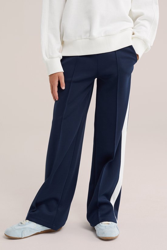 Model draagt een donkerblauwe wide leg broek met witte biezen en lichtblauwe sneakers.