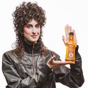 Persoon met donker krullend haar in leren jack houdt een fles Reuzel Grooming Tonic vast.