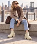 Model draagt beige Chelsea laarzen met blokhak, gecombineerd met jeans en een bruine sjaal.