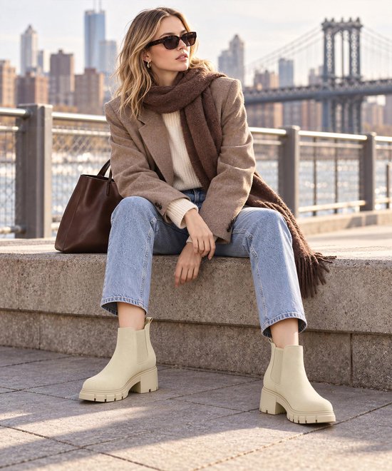 Model draagt beige Chelsea laarzen met blokhak, gecombineerd met jeans en een bruine sjaal.