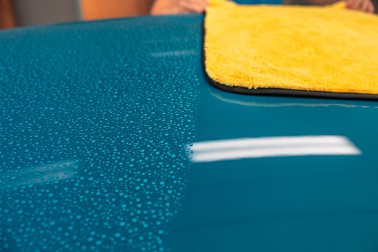 Een teal blauwe auto met waterdruppels aan de linkerkant en een felgeleende microvezel droogdoek aan de rechterkant.