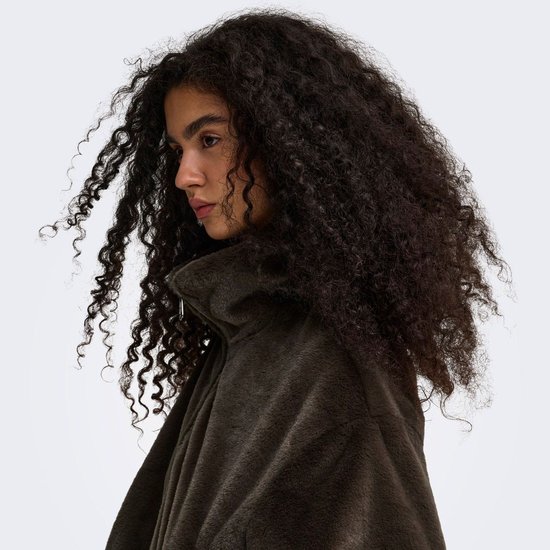 Close-up van een vrouw met krullend donker haar die het donkerbruine faux fur jack draagt en de kraag aanraakt.
