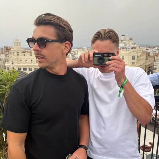Man met zonnebril en zwarte t-shirt kijkt weg, terwijl man achter hem met olijfgroene Phomo camera op een gebouw richt.