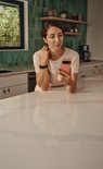 Vrouw in keuken kijkt op haar smartphone; draagt een zwarte fitnesstracker om haar pols.