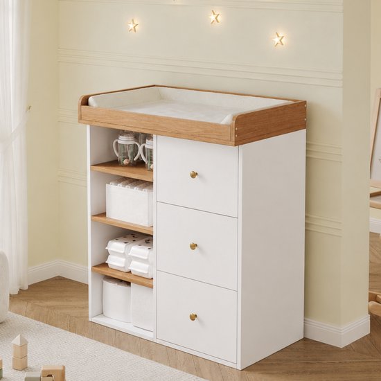 Wit-houten babycommode met aankleedkussen, drie lades en open zijkant met opbergmanden en flessen.