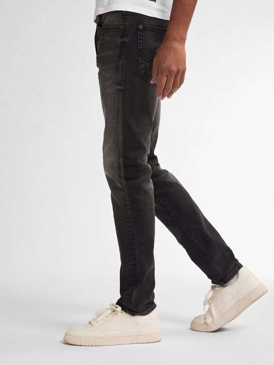 Model draagt zwarte slim fit jeans met vintage wassing, gecombineerd met beige sneakers in een zijwaartse pose.