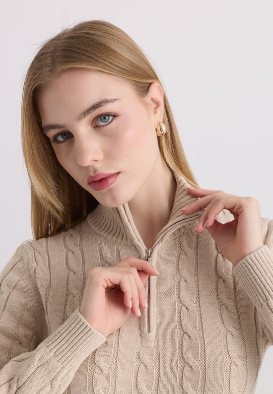 Close-up van een beige kabelgebreide trui met ritskraag, model houdt de kraag vast.