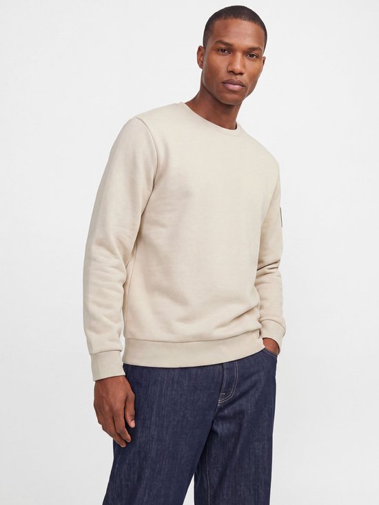 Man in een crème kleurige sweater en donkerblauwe jeans. De sweater heeft lange mouwen, boorden en een label op de mouw.