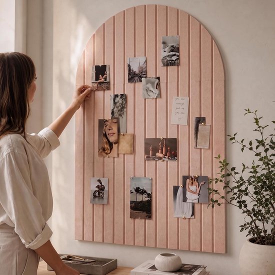 Vrouw in beige trui en linnen broek hangt foto's op aan een roze prikbord in de vorm van een boog.