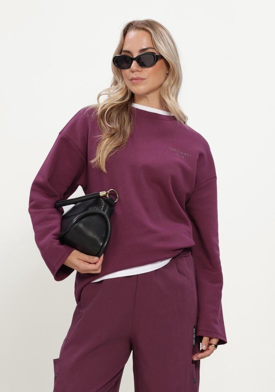 Model draagt bordeauxrode trui met "THE LABEL ALIX" opdruk, witte longsleeve en zwarte handtas.