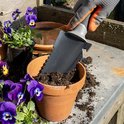 Hand met oranje-grijze handschoen gebruikt gekartelde zwarte spade om aarde in een bloempot te scheppen.