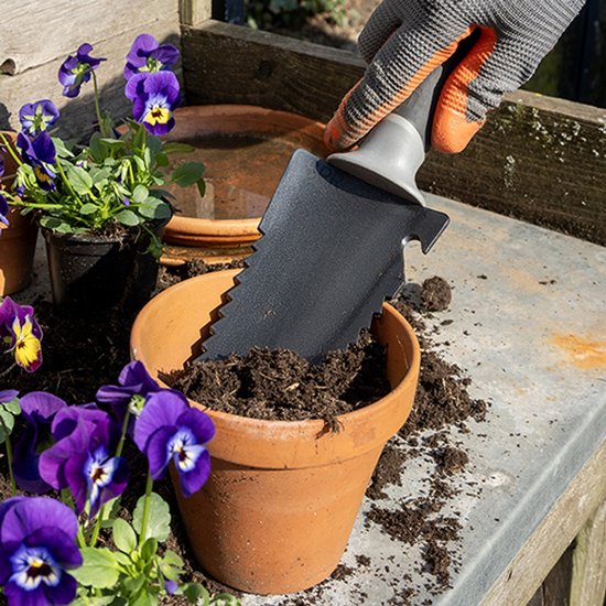 Hand met oranje-grijze handschoen gebruikt gekartelde zwarte spade om aarde in een bloempot te scheppen.