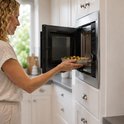 Vrouw met krullend blond haar doet een schaal met groenten in de Heinner inbouwmagnetron. De magnetron is ingebouwd in witte keukenkastjes.