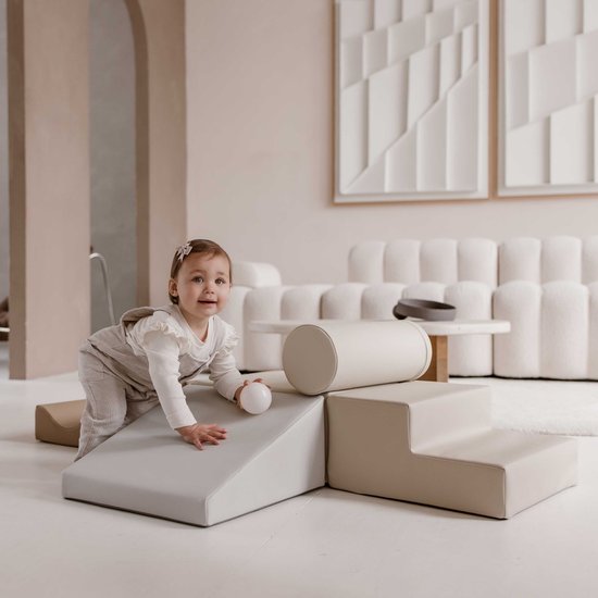 Een baby klimt op een beige en grijze speelset van schuimblokken in een woonkamer.