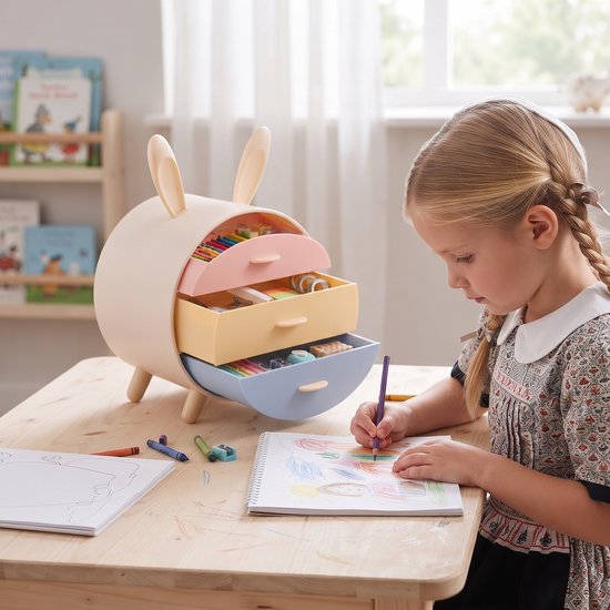Een meisje met blonde vlechtjes kleurt in een schetsboek aan een houten tafel, naast een opberger in konijnenoren-stijl met pastelkleurige lades.