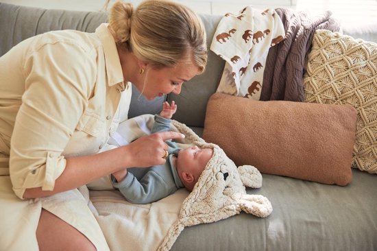 Een baby in een beige wikkeldeken met konijnenoren ligt op een bank bij een vrouw.