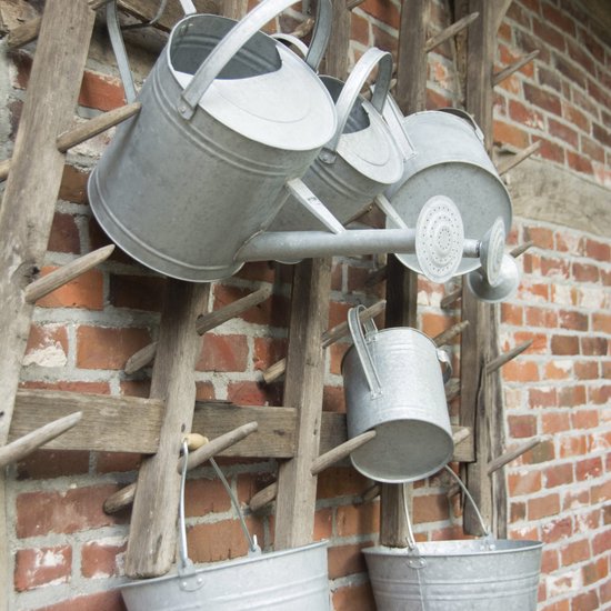 Drie zinken gieters en twee emmers hangen aan een houten rek voor een bakstenen muur.