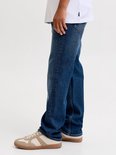 Zijaanzicht van een jongen in witte t-shirt en blauwe jeans met bruine sneakers.