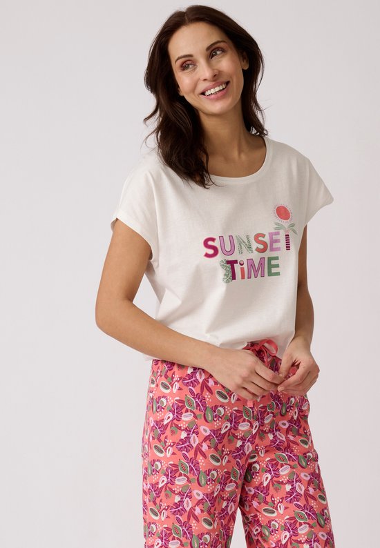 Vrolijke vrouw in witte top met 'SUNSET TIME' opdruk en roze broek met tropische print.