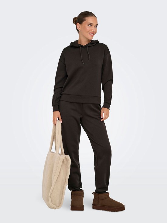 Model met donkerbruine joggingbroek en hoodie, draagt bruine laarsjes en draagt een beige teddy shopper.