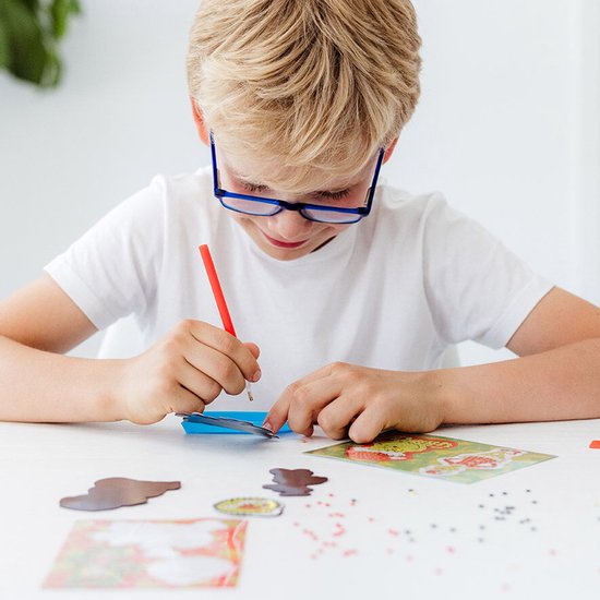 Garçon avec des lunettes créant un aimant Peinture Diamant du Grinch, plaçant les strass avec un stylo rouge.