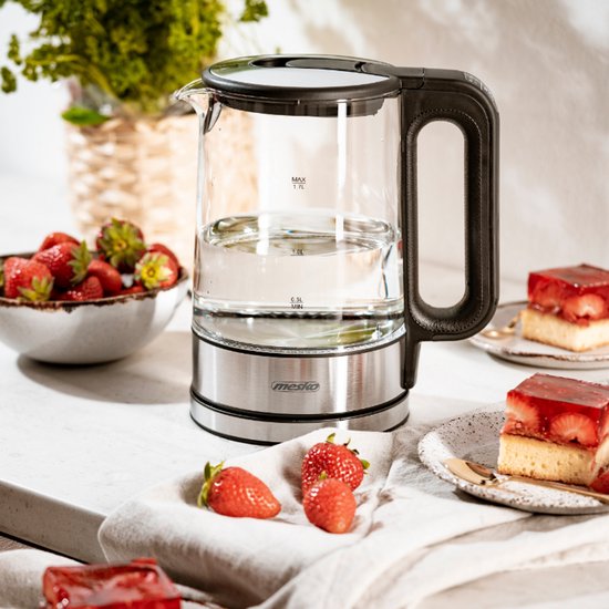 Glazen waterkoker met zwart handvat en RVS voet, gevuld met water, op een tafel met aardbeien en gebak.