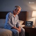 Oudere man in pyjama zit op bed naast een digitale klok met "DINSDAG NACHT 11:32 20 JANUARI 2026".