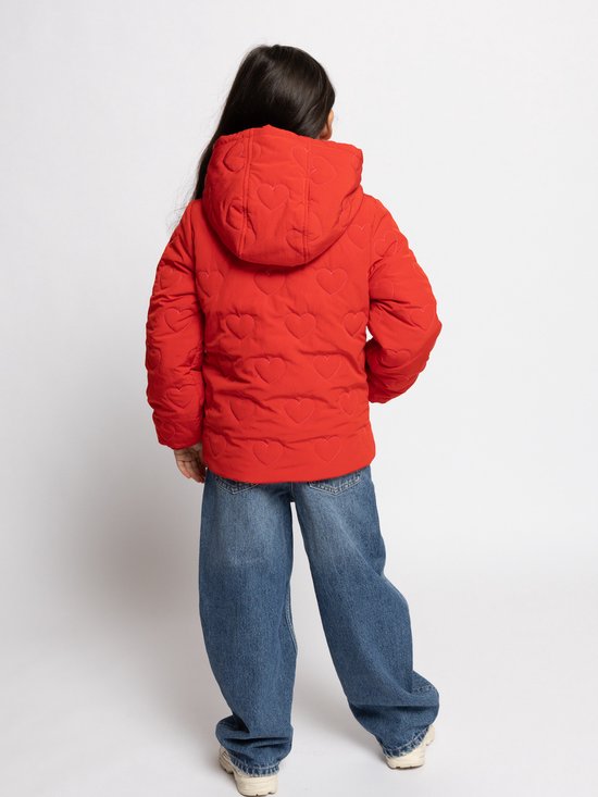 Meisje met lange donkere haren, draagt een rode jas met hartjes en spijkerbroek.