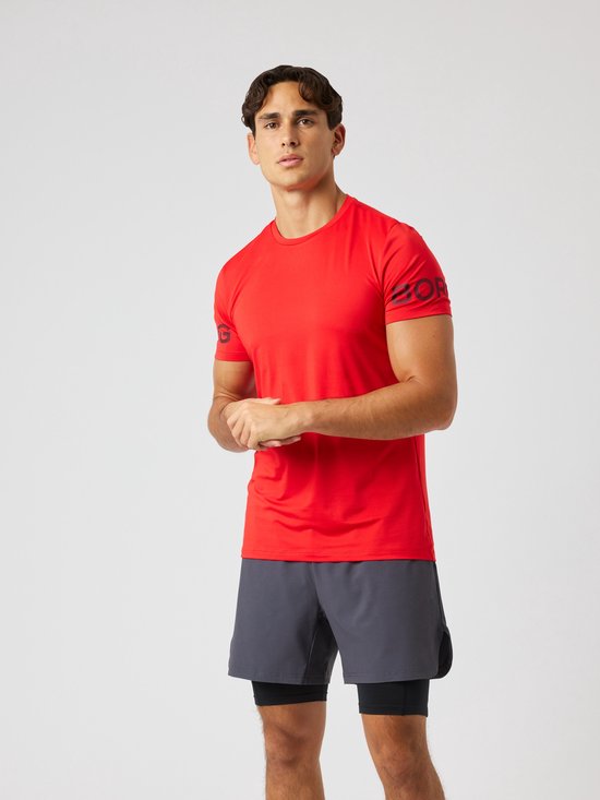 Model staat frontaal in een felrood sportshirt met zwarte opdruk op de mouwen. Draagt een grijze sportshort. Witte sneakers en sokken.