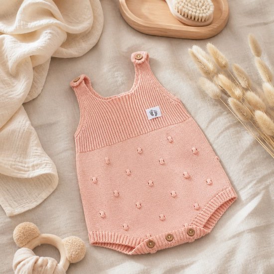 Roze gebreid babyrompertje met knoopjes aan de schouders en subtiel stipjespatroon.