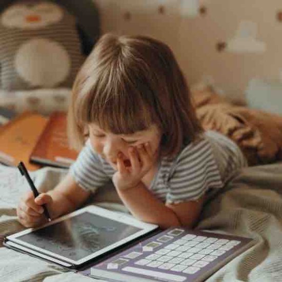 Een kind met bruin haar, liggend op een bed, tekent op een tablet met een stylus.