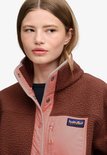 Close-up van de kraag en het logo van een bruine fleece jas met roze panelen.