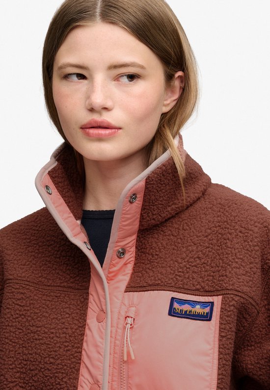 Close-up van de kraag en het logo van een bruine fleece jas met roze panelen.