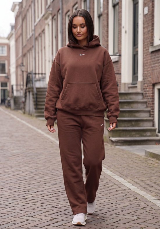 Vrouw loopt door een straat in een bruine Nike joggingbroek en hoodie. De outfit heeft een klein wit Nike logo.