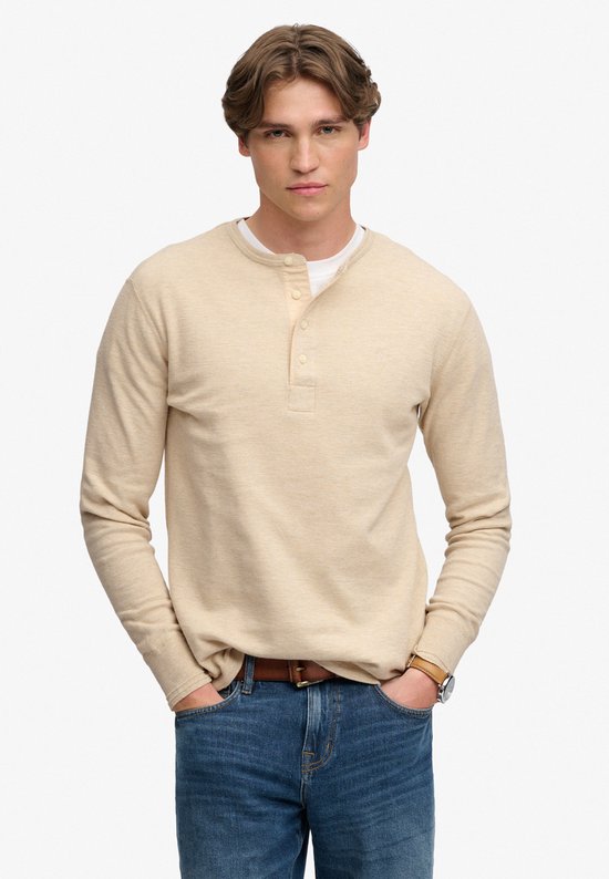 Model draagt een beige waffle grandad-top met lange mouwen en knoopsluiting, gecombineerd met een blauwe jeans.