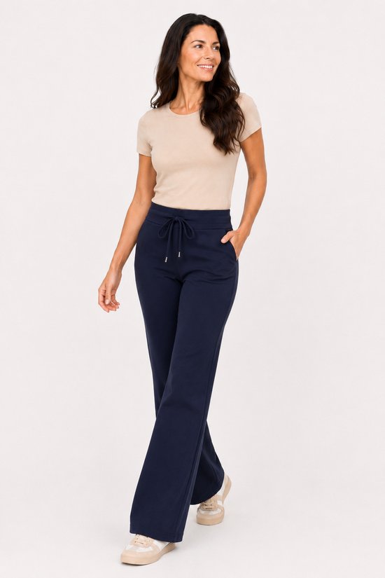 Vrouw in een beige top en marineblauwe flared broek met trekkoord, staand met de hand in de zak.